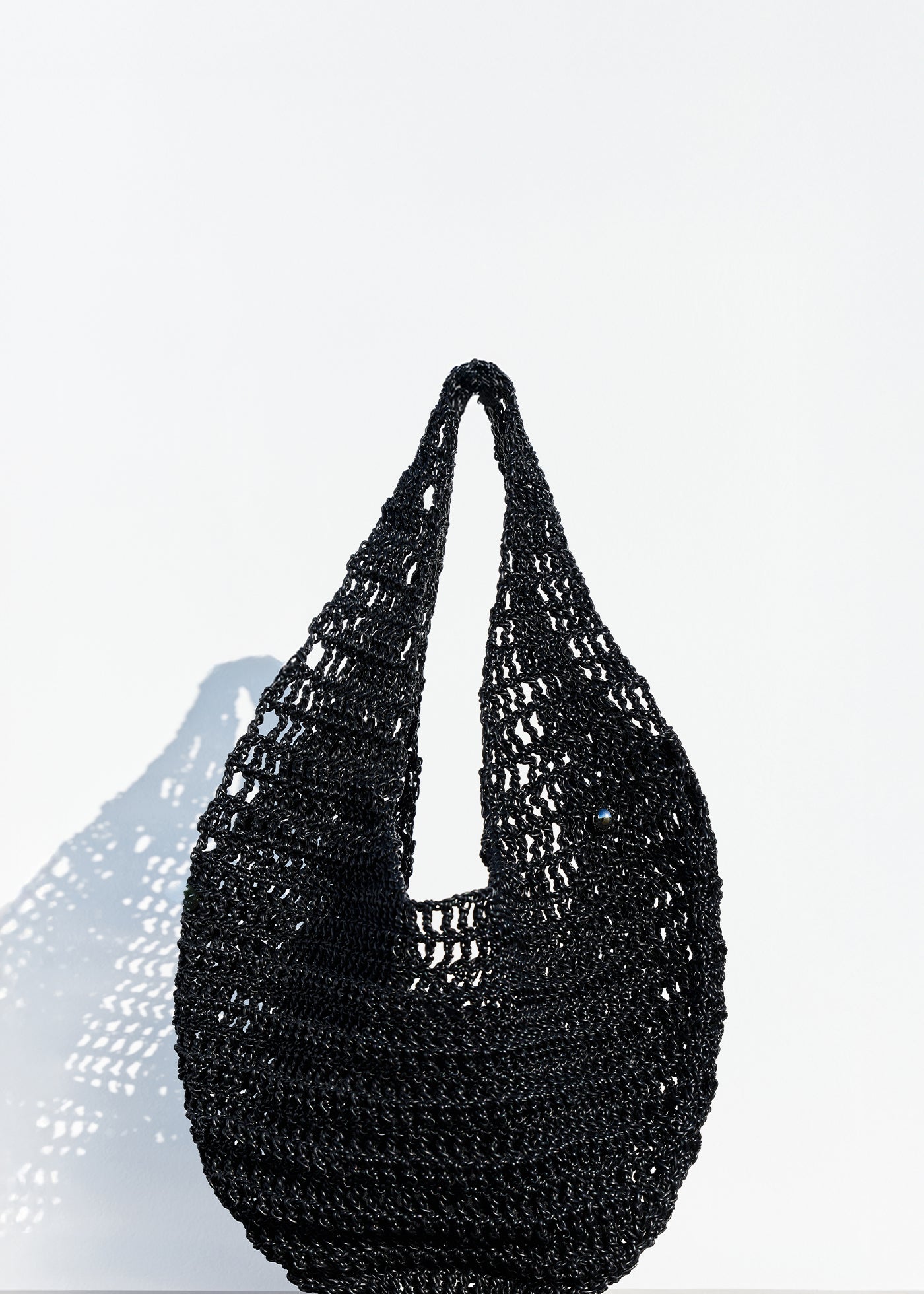 The Market Net Bag, a black, woven crochet-style handbag with a single looped handle, sits on a light wooden surface against a plain white background.