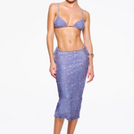 A woman stands against a white background wearing The C Skirt, a high-waisted, knee-length skirt with a purple floral print and side ties. The bias cut silhouette flatters her figure and she pairs it with a coordinating bikini-style top, white heeled sandals, and minimal accessories.
