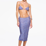 A woman stands against a white background wearing The C Skirt, a high-waisted, knee-length skirt with a purple floral print and side ties. The bias cut silhouette flatters her figure and she pairs it with a coordinating bikini-style top, white heeled sandals, and minimal accessories.