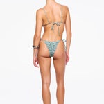 A woman stands facing away, wearing a light green patterned recycled nylon swimwear bikini and sandals, with her hair tied back. The bikini features beaded tie side straps on both the top and the zebra sage Praia Bottom. The background is plain white.