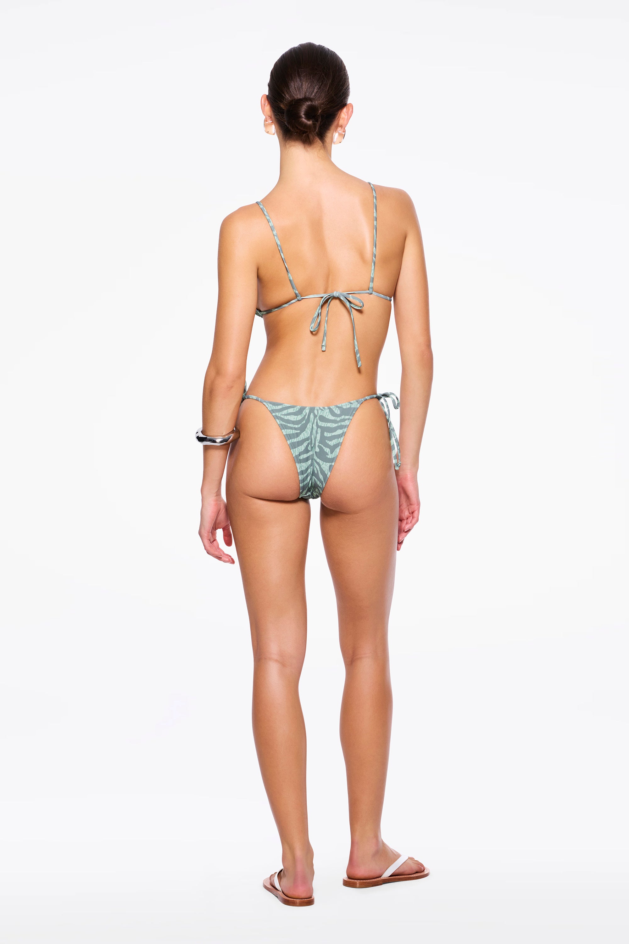 A woman stands facing away, wearing a light green patterned recycled nylon swimwear bikini and sandals, with her hair tied back. The bikini features beaded tie side straps on both the top and the zebra sage Praia Bottom. The background is plain white.