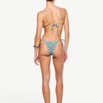 A woman stands facing away, wearing a light green patterned recycled nylon swimwear bikini and sandals, with her hair tied back. The bikini features beaded tie side straps on both the top and the zebra sage Praia Bottom. The background is plain white.