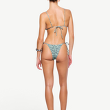 A woman stands facing away, wearing a light green patterned recycled nylon swimwear bikini and sandals, with her hair tied back. The bikini features beaded tie side straps on both the top and the zebra sage Praia Bottom. The background is plain white.