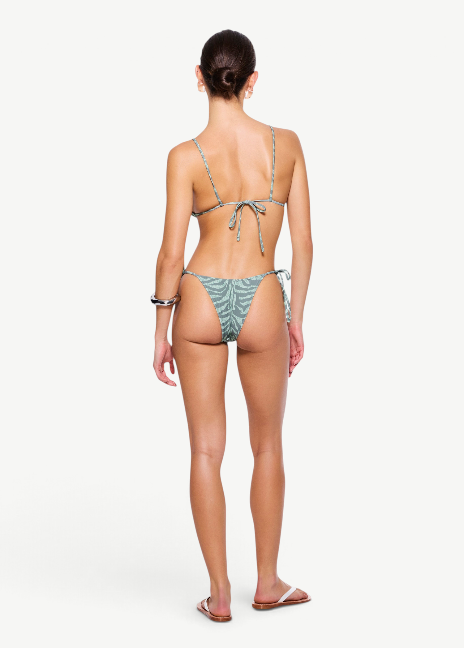 A woman stands facing away, wearing a light green patterned recycled nylon swimwear bikini and sandals, with her hair tied back. The bikini features beaded tie side straps on both the top and the zebra sage Praia Bottom. The background is plain white.