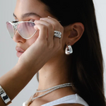 A woman with long dark hair wears silver sunglasses and the Jordyn Hoops in Rhodium—chunky hoop earrings with a pierced back closure. She pairs them with a white tank top and holds her sunglasses near her eyes, gazing to the side against a plain background.