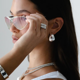 A woman with long dark hair wears silver sunglasses and the Jordyn Hoops in Rhodium—chunky hoop earrings with a pierced back closure. She pairs them with a white tank top and holds her sunglasses near her eyes, gazing to the side against a plain background.
