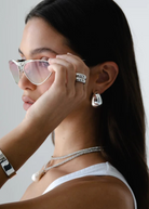 A woman with long dark hair wears silver sunglasses and the Jordyn Hoops in Rhodium—chunky hoop earrings with a pierced back closure. She pairs them with a white tank top and holds her sunglasses near her eyes, gazing to the side against a plain background.