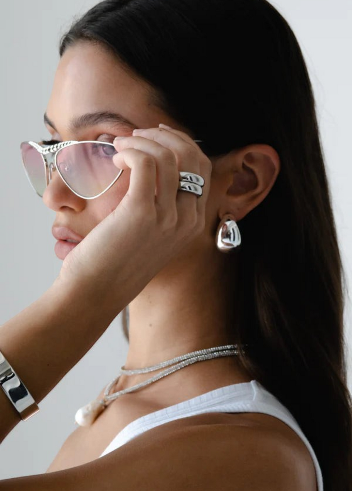 A woman with long dark hair wears silver sunglasses and the Jordyn Hoops in Rhodium—chunky hoop earrings with a pierced back closure. She pairs them with a white tank top and holds her sunglasses near her eyes, gazing to the side against a plain background.